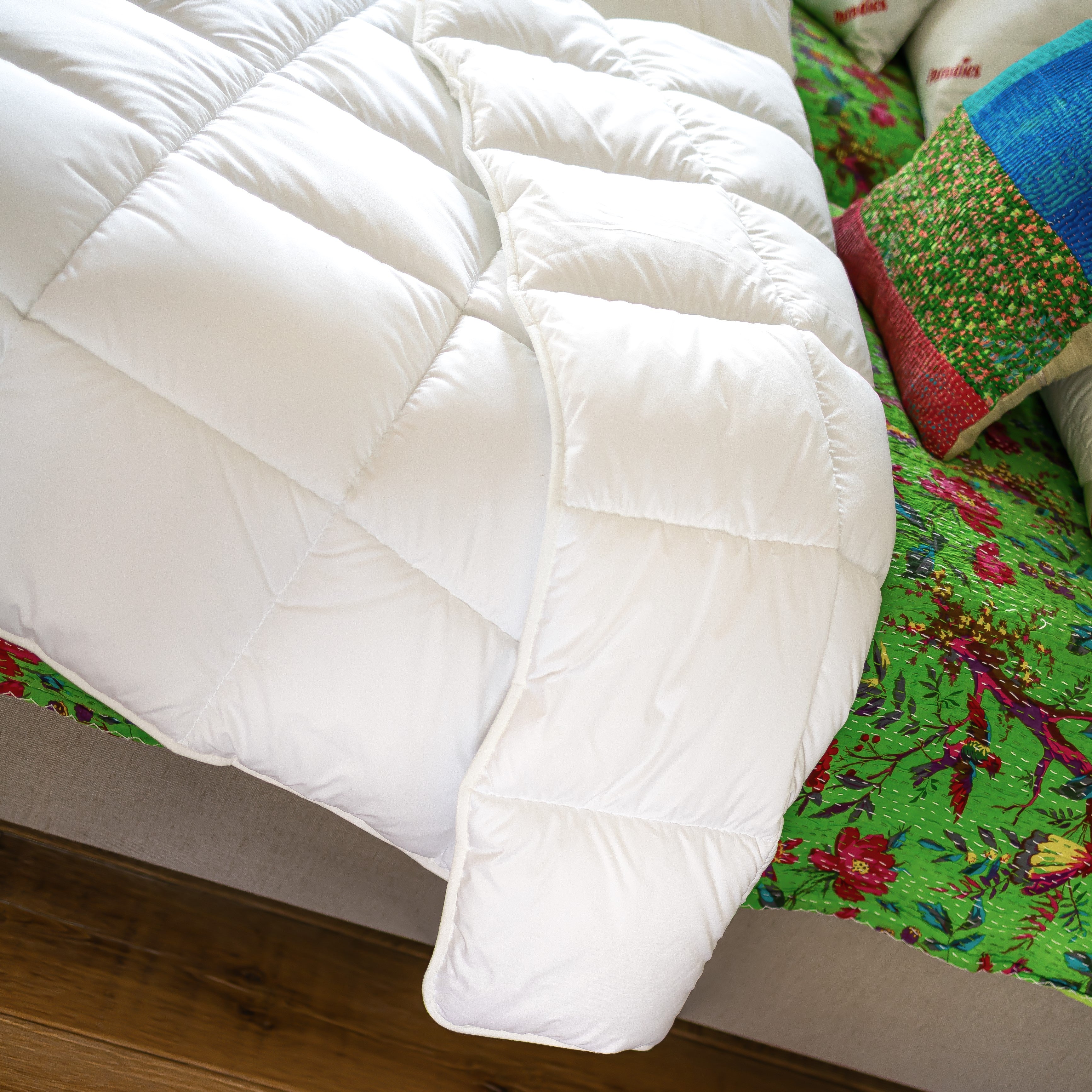 Weiße Steppdecke liegt gefaltet auf Bett mit grün-rotem Blumenmuster und buntem Kissen.