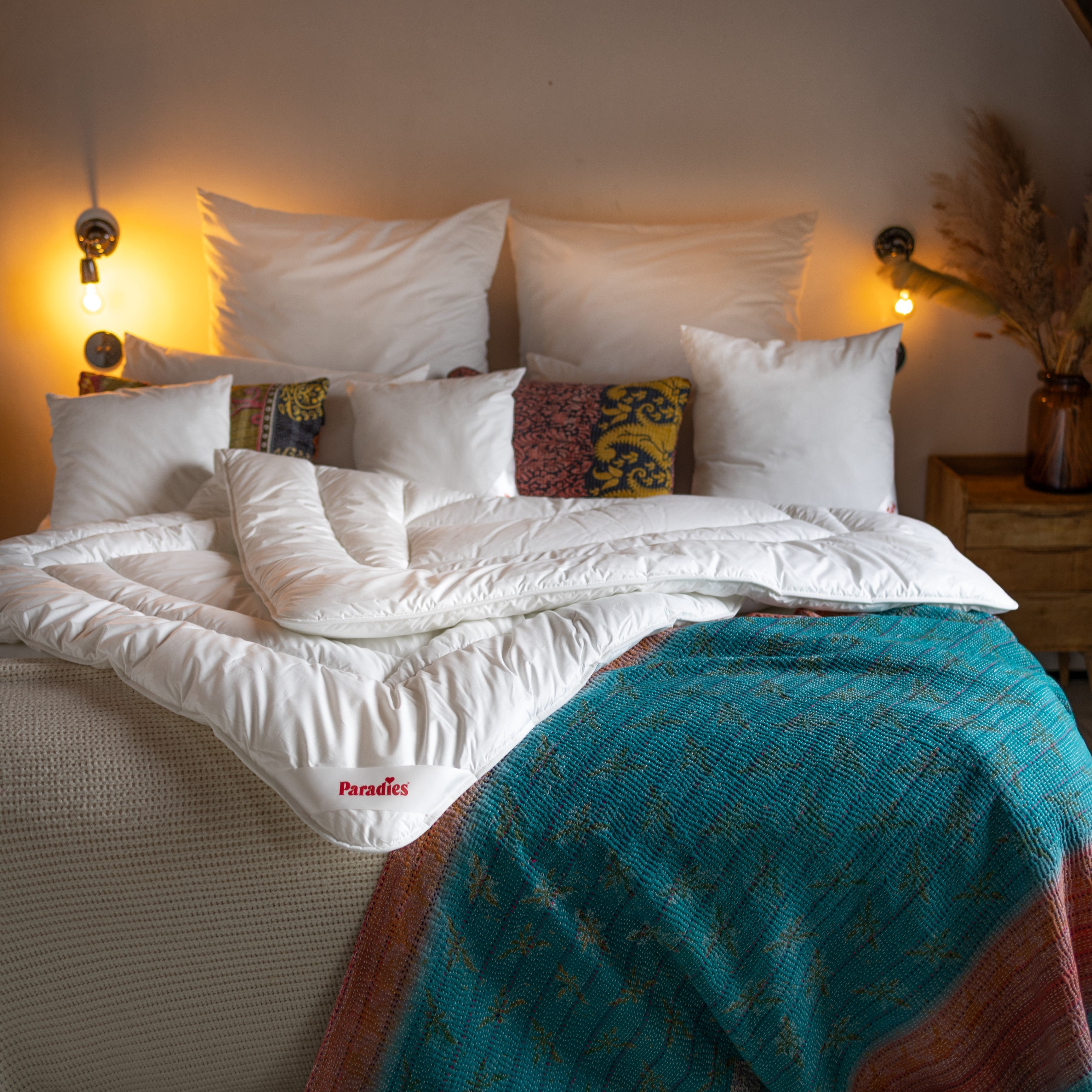 Weiße gefaltete Ganzjahresdecke mit rotem 'Paradies'-Logo auf einem Bett mit mehreren weißen und gemusterten Kissen, zwei Wandlampen leuchten.