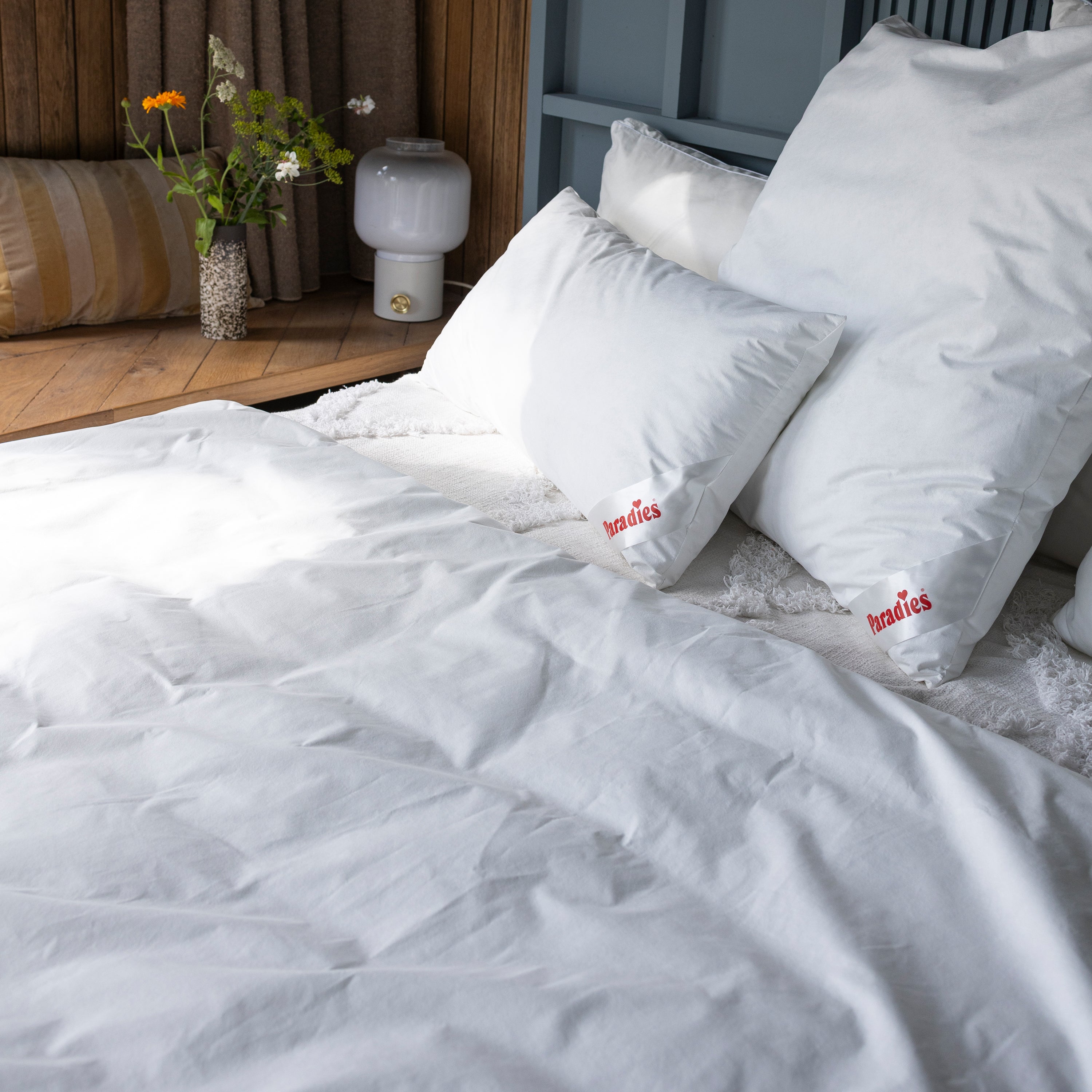 Zwei weiße Paradies Softy Antiallergo Kissen auf einem Bett mit Antiallergo Bettdecke. Im Hintergrund eine Holzablage mit Kissen, Vase und Nachttisch-Lampe