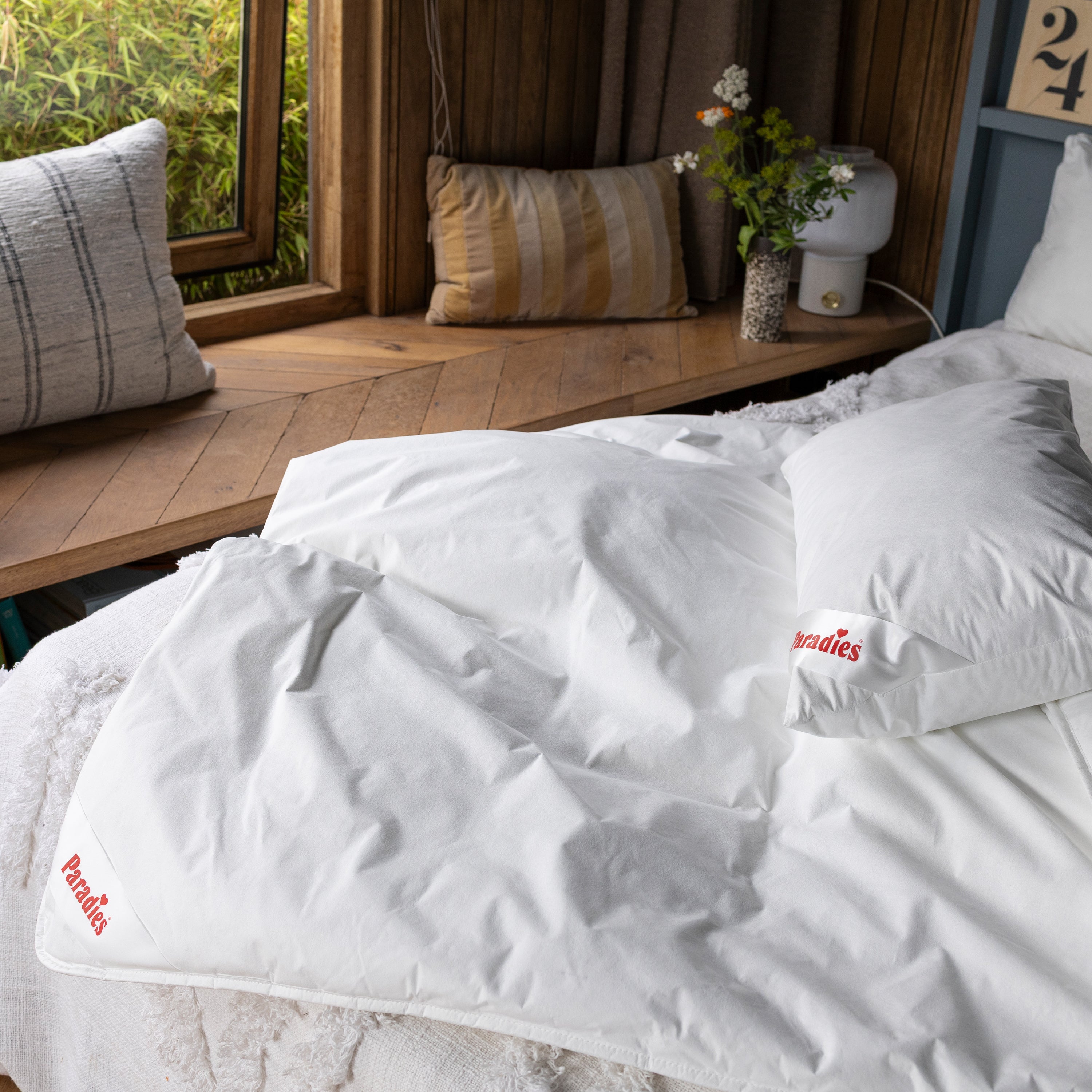 Weiße Paradies Antiallergo Bettdecke mit einem Softy Antiallergo Kissen auf einem Bett. Im Hintergrund eine Holzablage mit Kissen, Vase und Nachttisch-Lampe