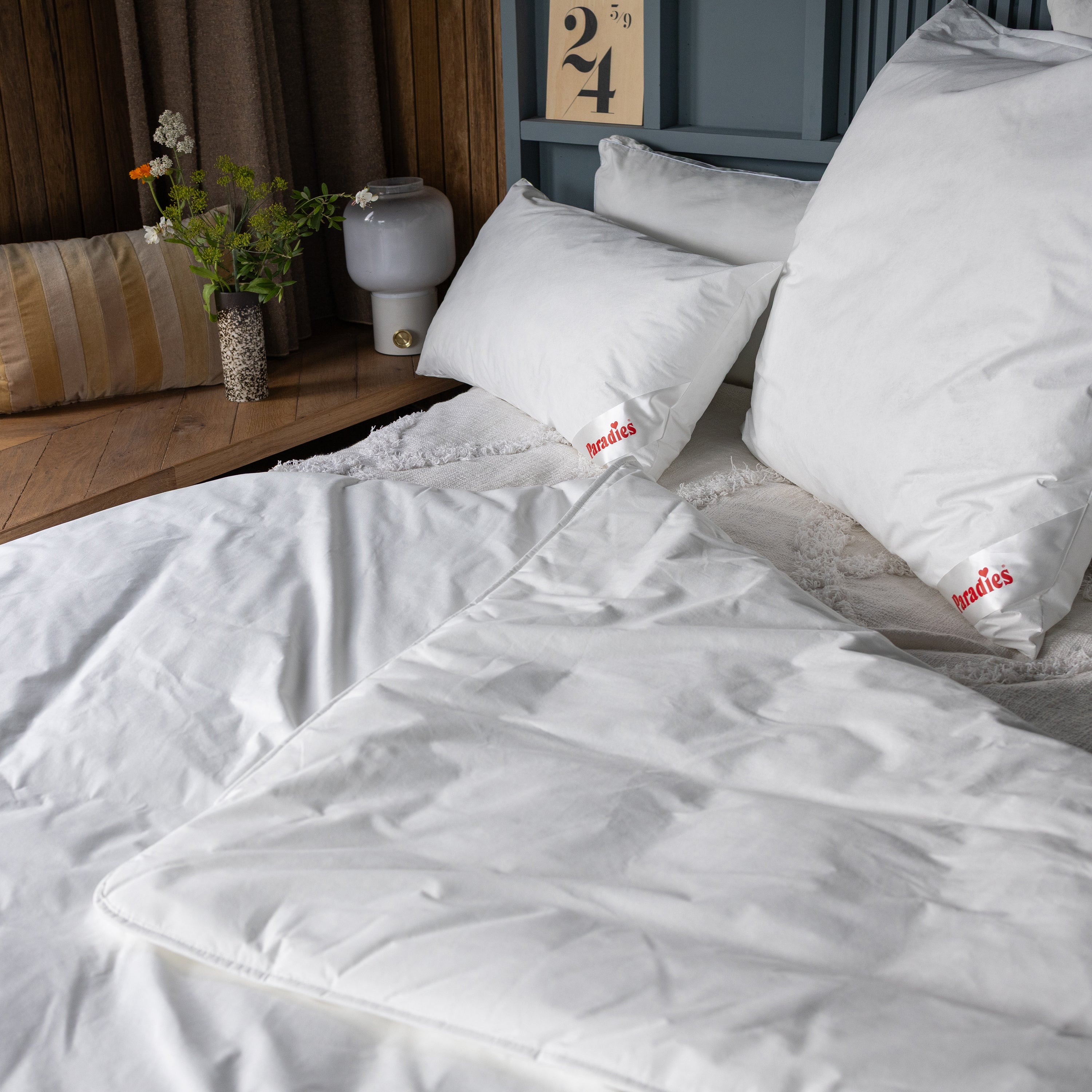 Zwei weiße Paradies Softy Antiallergo Kissen auf einem Bett mit Antiallergo Bettdecke. Im Hintergrund eine Holzablage mit Kissen, Vase und Nachttisch-Lampe