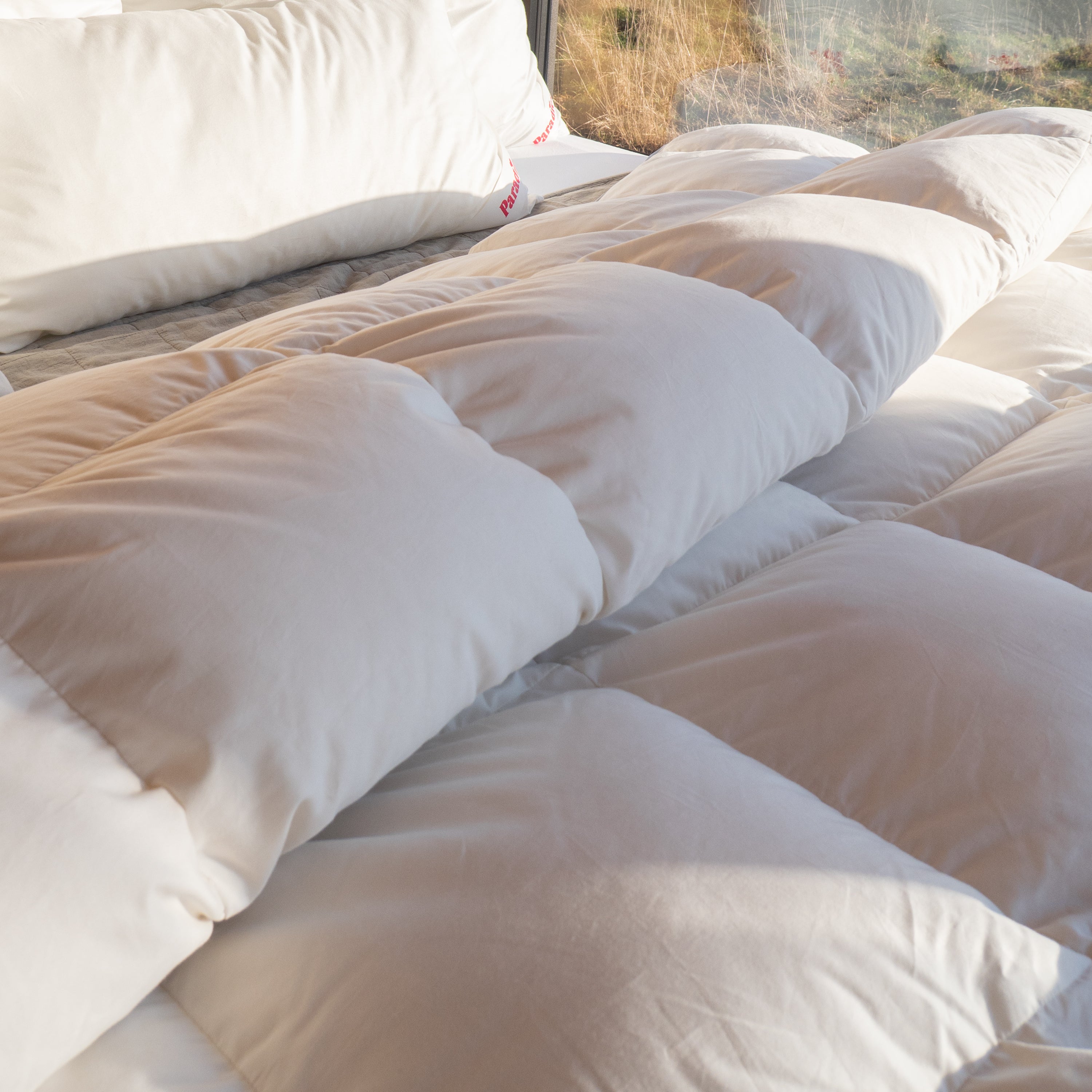 Weiße Paradies Winterdaunendecke mit voluminösen Kammern, die gleichmäßig gefüllt und weich erscheinen, liegt zusammen mit zwei Kissen auf Bett vor einem Fenster