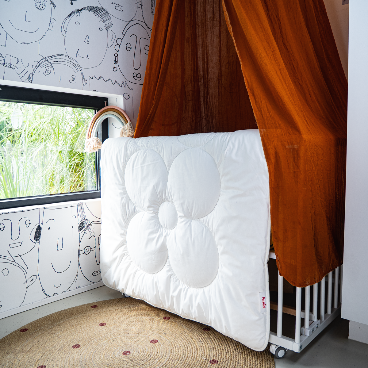 Weiße Kinderbettdecke mit großer Blütenform, 135x100 cm, hängt an einem weißen Kinderbett mit braunem Himmel in einem Kinderzimmer mit gezeichneten Gesichtern an der Wand