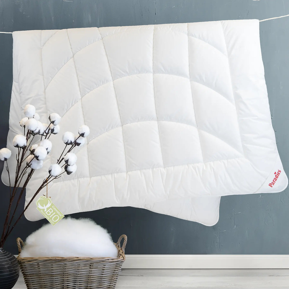 Paradies Premium Bettdecke Runa Bio Winterdecke hängt an Wäscheleine mit einer Baumwollpflanze und einem Korb gefüllt mit Faservlies