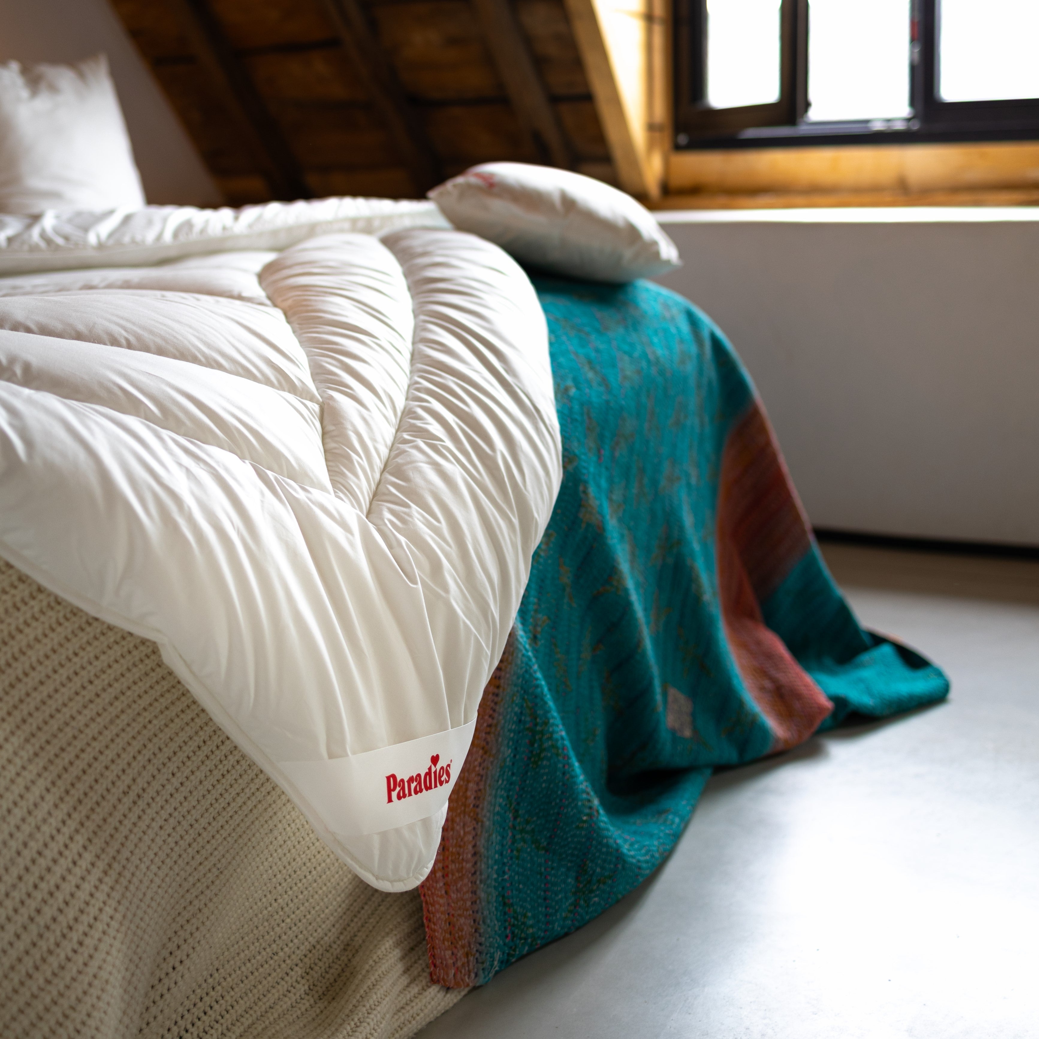 Weiße Avera Ganzjahresdecke mit Steppmuster und rotem 'Paradies'-Label hängt von einem Bett mit buntem Überwurf und Kissen in einem Raum mit Dachschräge und Fenster.