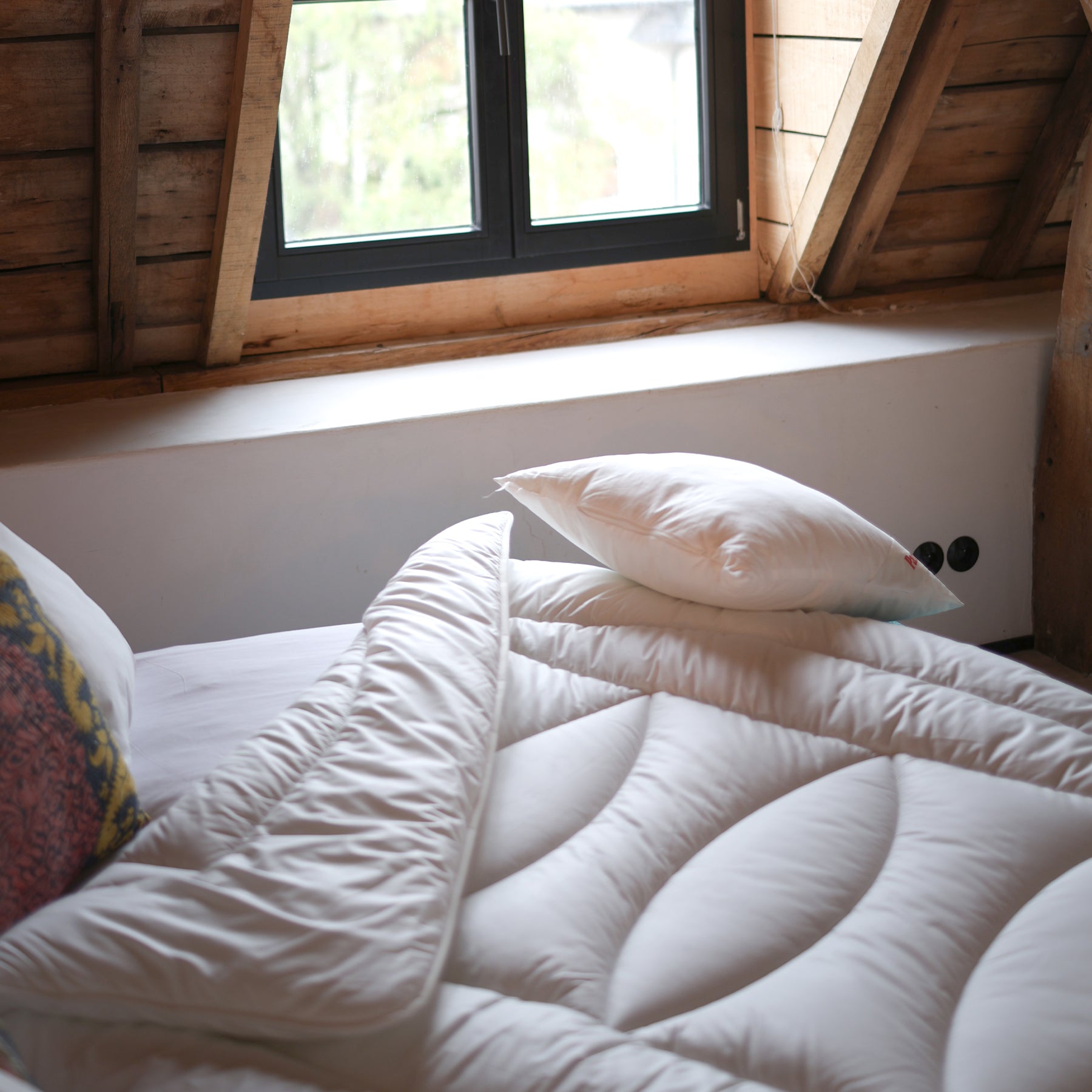 Bett mit weißer Avera Bettdecke und Kissen unter einem Fenster in einem Raum mit Dachschräge.