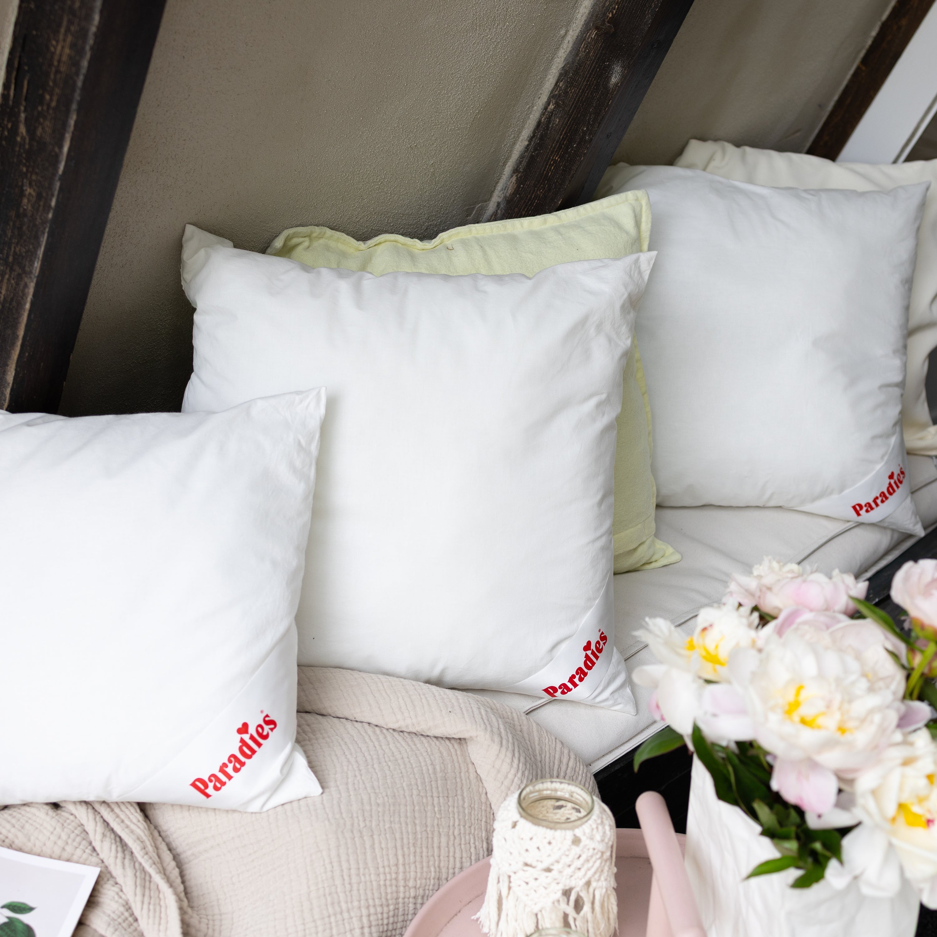 Drei weiße Paradies Kuschelkissen auf einer Bank neben einem Blumenstrauß und einer Kerze