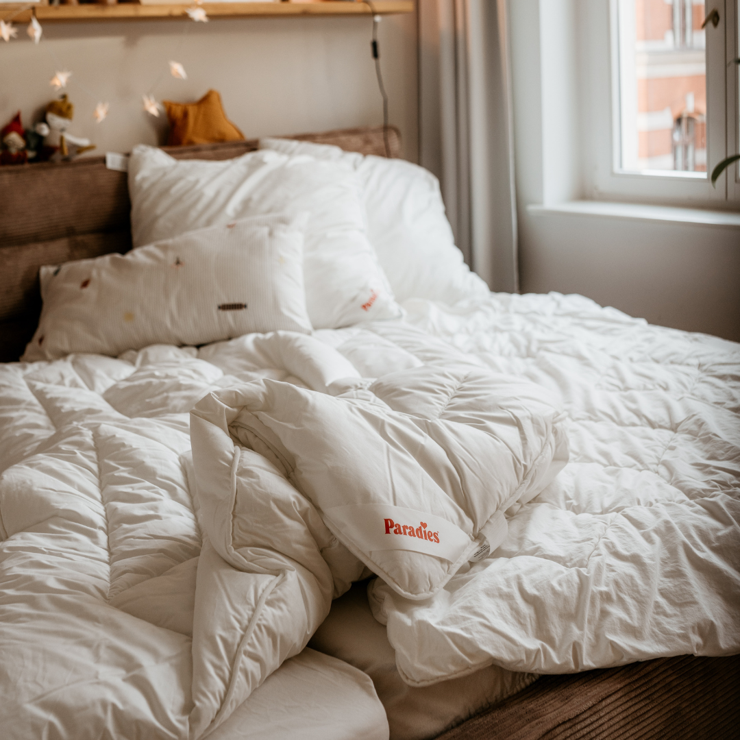 Weißes, gefaltetes Winterbett mit gestepptem Muster und Paradies Schriftzug auf einem Bett in einem hellen Schlafzimmer