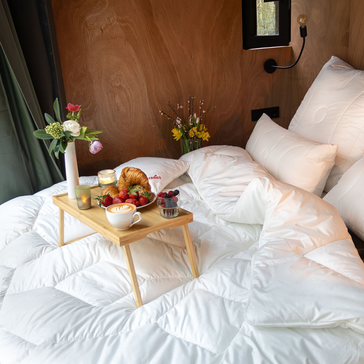 Holztablett mit Croissant, Beeren, Kaffee und Kerze auf weißer Bettdecke in gemütlichem Schlafzimmer mit Holzwand