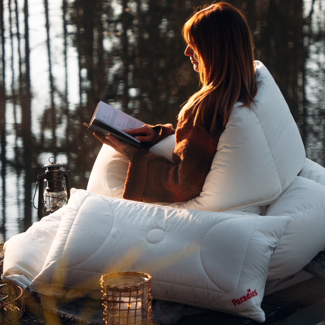 Weiches, weißes Kissen mit Steppnähten und rotem Schriftzug "Paradies" liegt vor einer Person, die in einer Decke eingehüllt ein Buch liest.